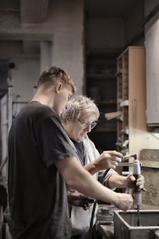 A young apprentice works with a senior craftsman in a workshop, focusing on handwork.