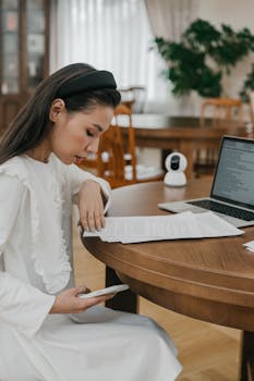 Focused Asian woman studying with laptop and smartphone at home, emphasizing online learning and education.