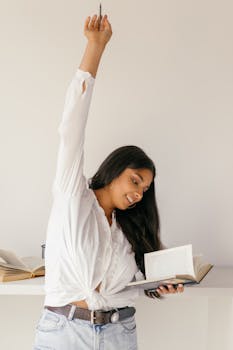 Smiling young woman indoors, raising her hand in celebration while studying a book.